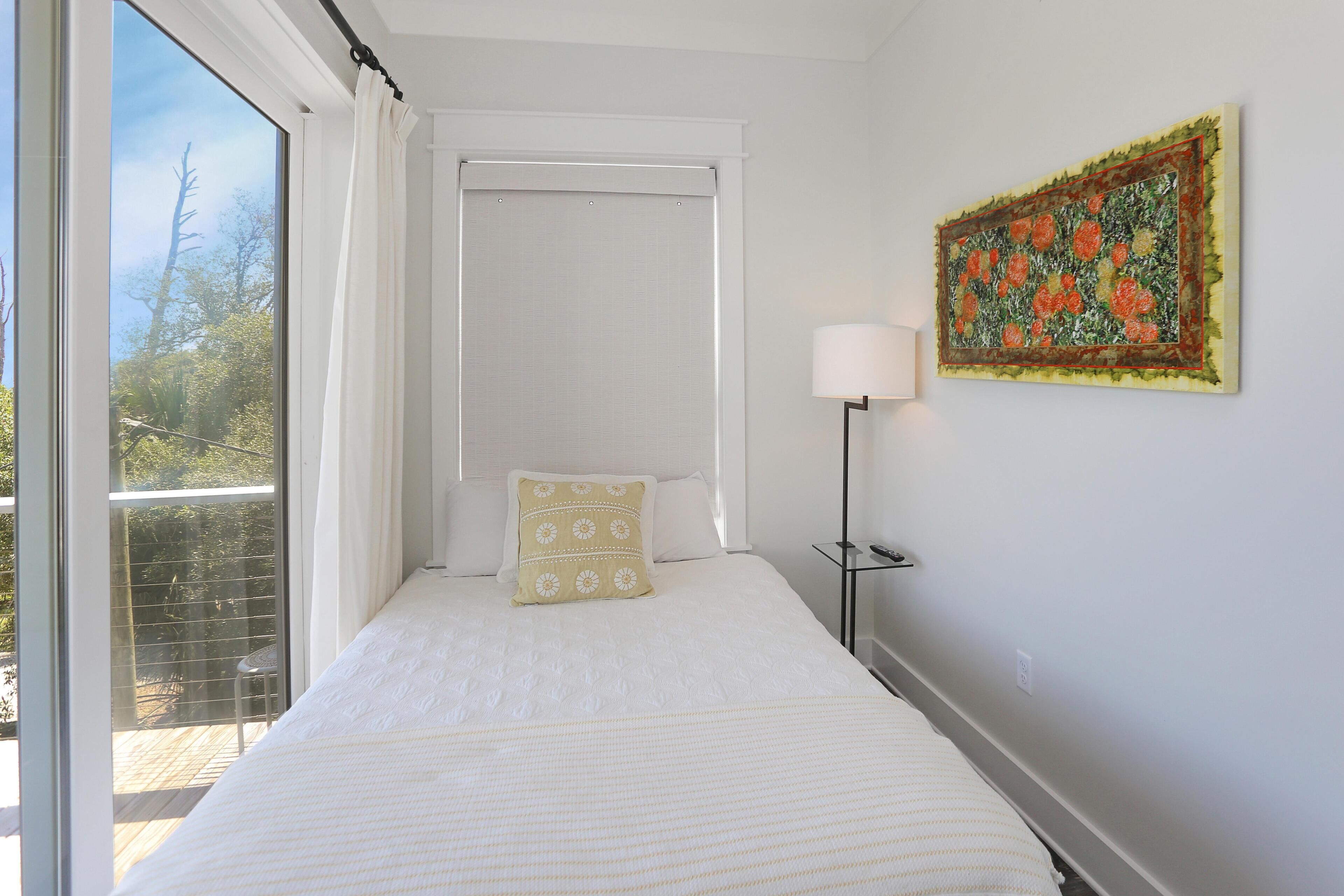 A bedroom with a large window, a bed with white bedding, a bedside table with a lamp, and a wall-mounted artwork.