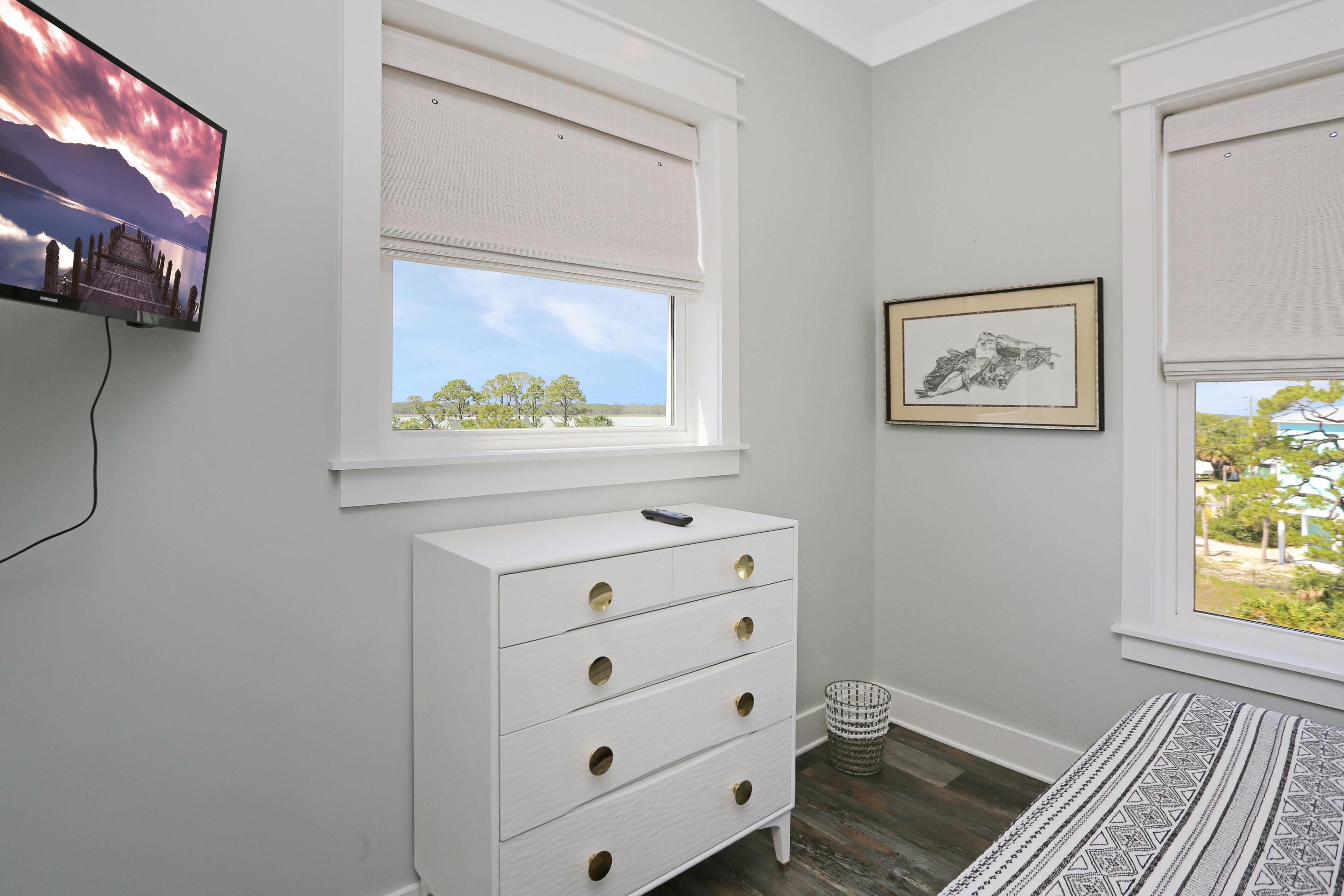 A bedroom with a white dresser, a television mounted on the wall, and a window with a view of trees.