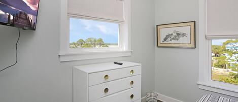 A bedroom with a white dresser, a television mounted on the wall, and a window with a view of trees.