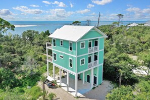 Un edificio turchese a più piani con balconi, immerso nel verde e con una spiaggia sullo sfondo.