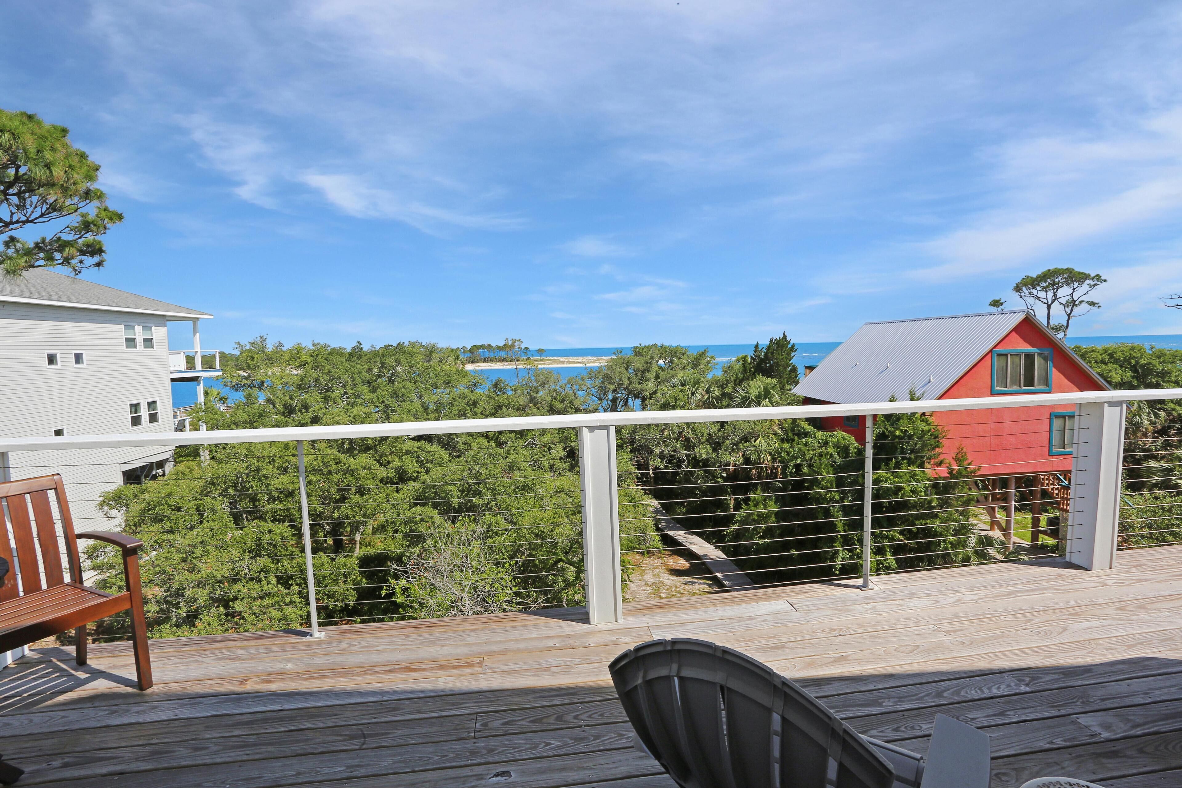 A wooden deck with a bench and a chair, overlooking a beach and a red house.