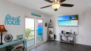 A beach-themed living room with a ceiling fan, a TV displaying a beach scene, a glass coffee table, and a door leading to a balcony.