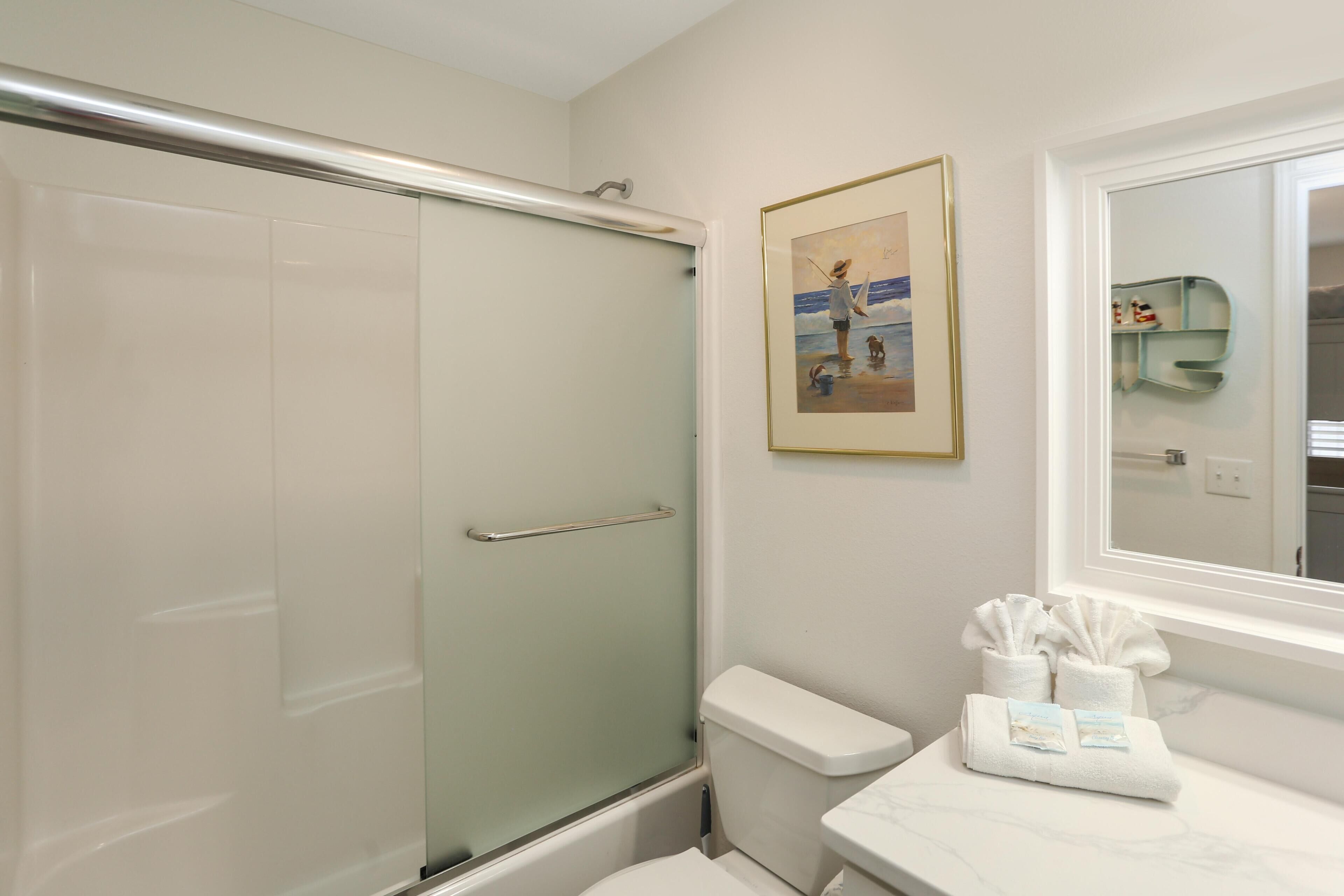 A bathroom with a glass shower enclosure, a toilet, a marble sink with toiletries, and a framed picture on the wall.