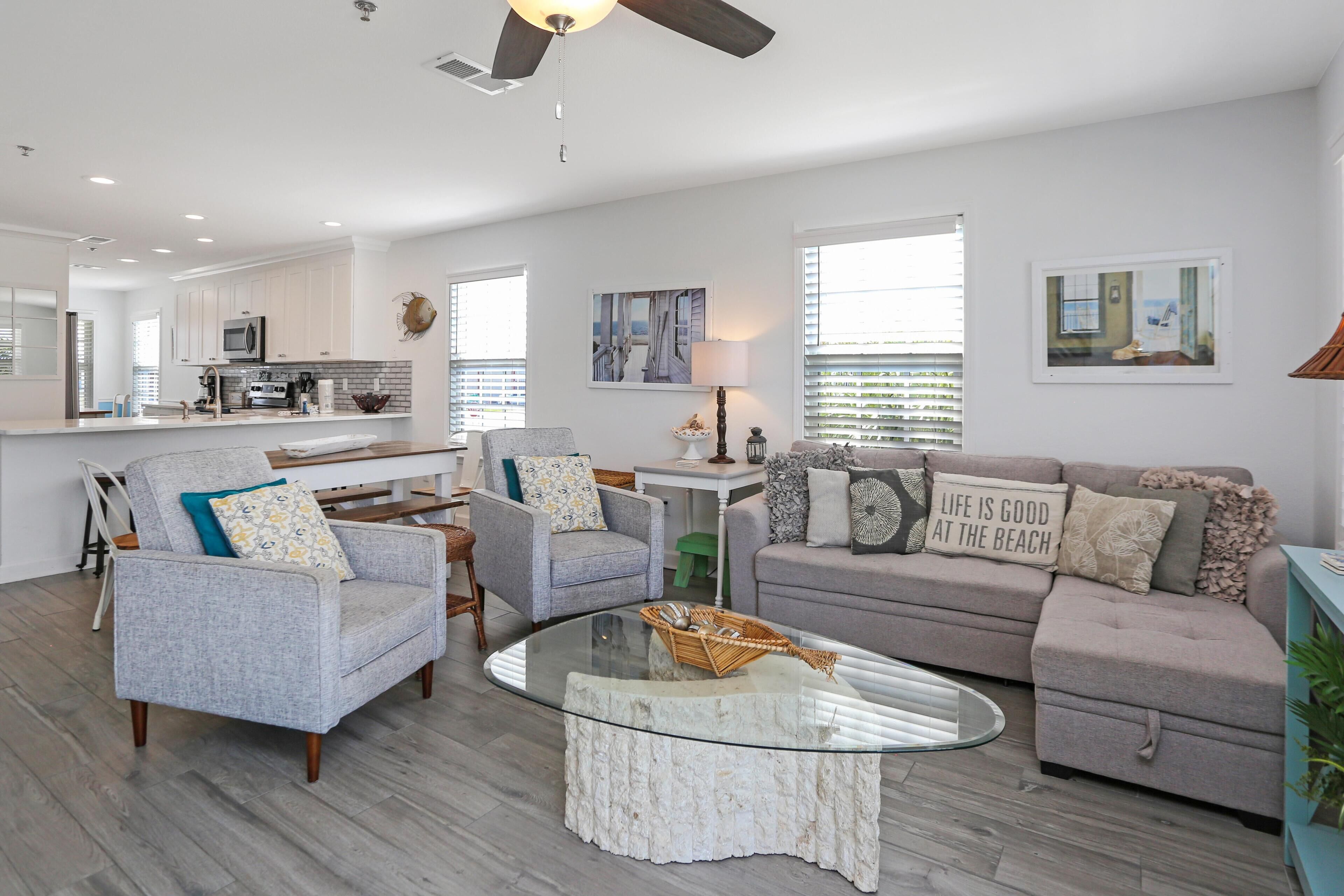 A modern living room with a glass coffee table, grey sofas, and a kitchen in the background.