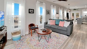 A spacious living room with a grey sofa, a coffee table, and a large window with white curtains. - Hesed Beach Cottage (Port St. Joe)