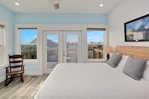 A bedroom with a large bed, a wooden chair, a painting, and a view through the glass doors. - Hesed Beach Cottage (Port St. Joe)