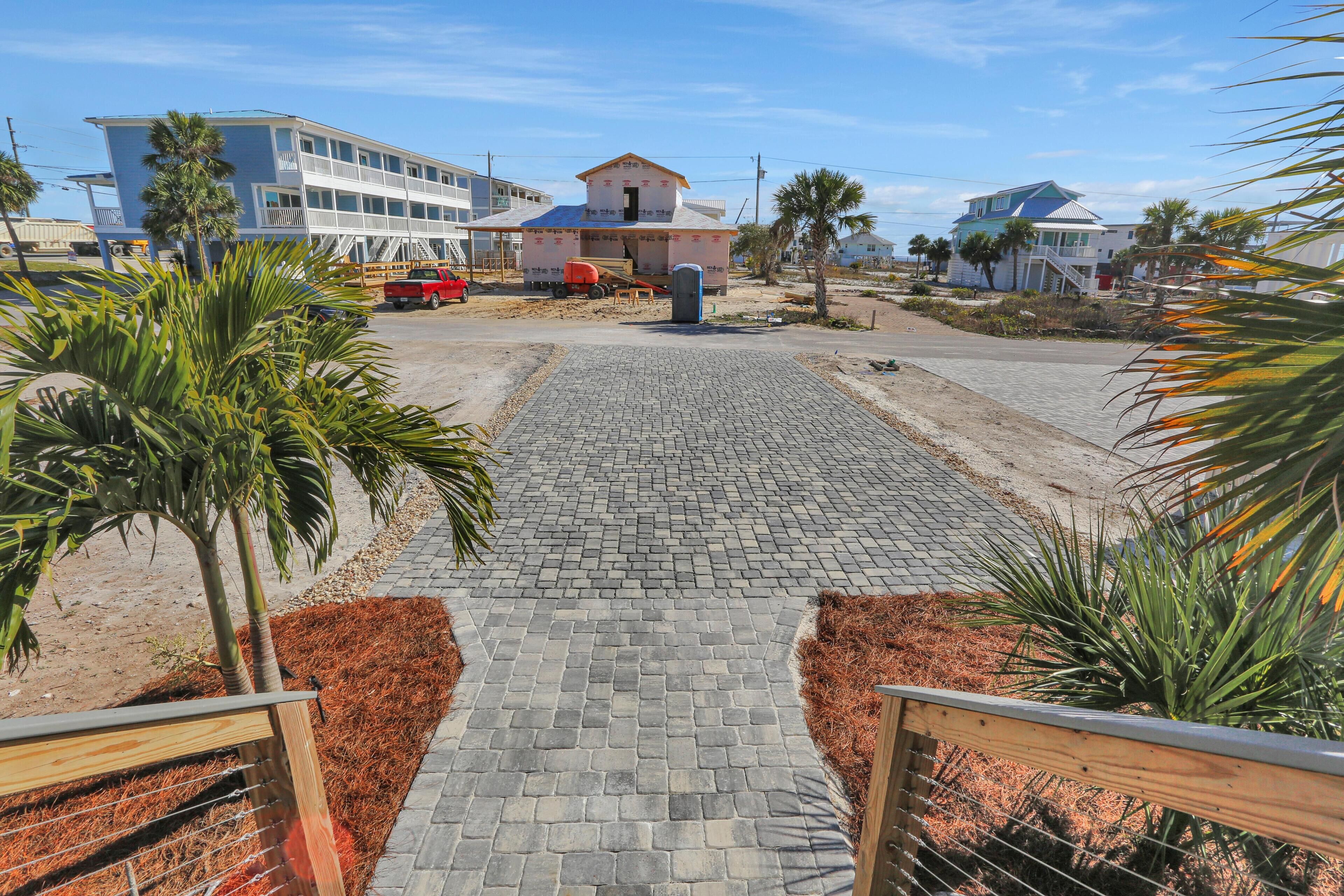 A paved pathway leading to a building with a balcony, surrounded by palm trees and a sandy area.