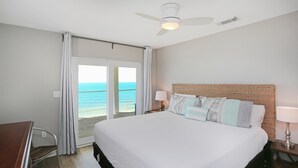 Habitación de hotel con cama, escritorio, silla, ventilador de techo y vista al océano a través de una puerta corrediza.