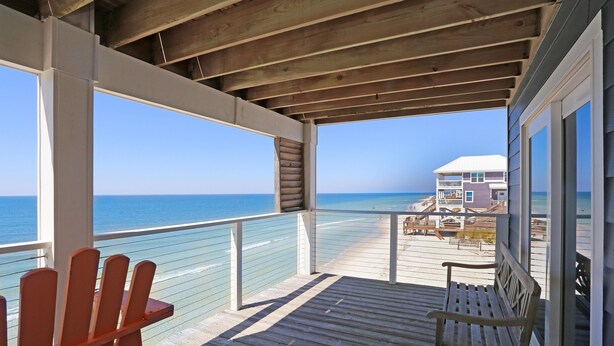 Una terraza de madera con un banco, con vistas a una playa y una casa al fondo.