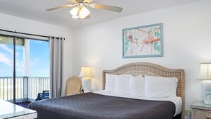 A hotel room with a bed, a ceiling fan, a window with curtains, a painting on the wall, and a nightstand with a lamp. - Flamingo Dunes (Port St. Joe)