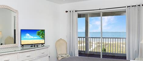 A bedroom with a large bed, a television on a dresser, a ceiling fan, and a balcony with a view of the beach.