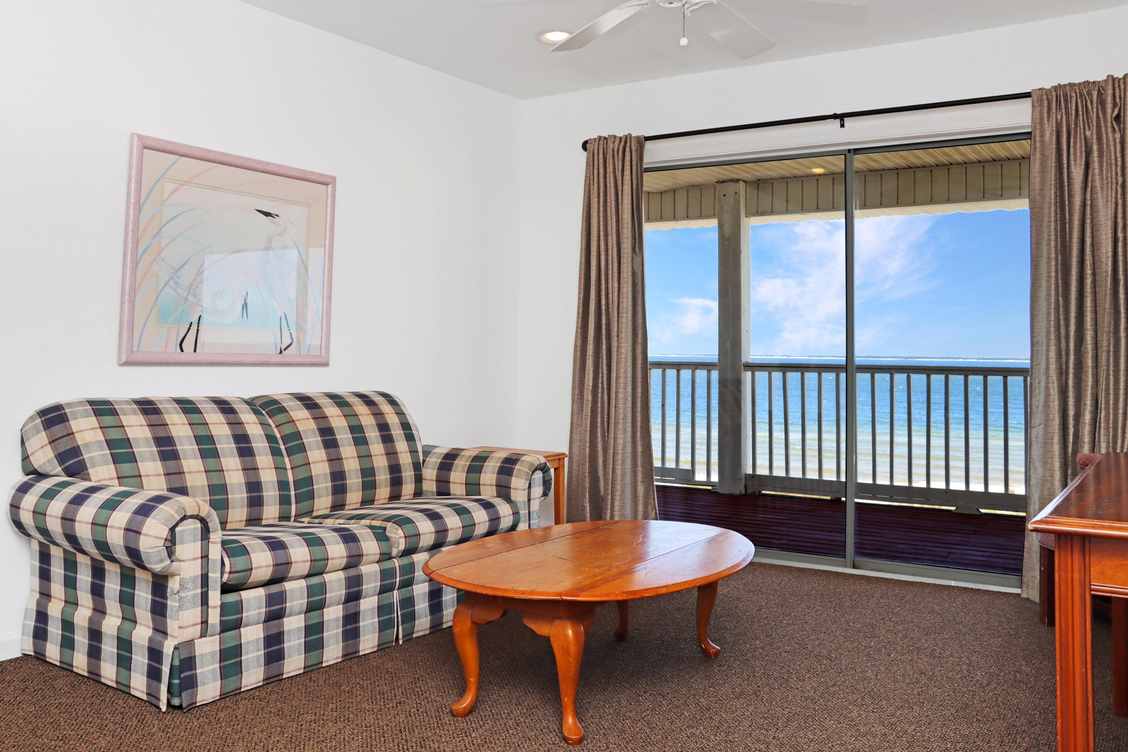 A living room with a plaid sofa, a wooden coffee table, a painting on the wall, and a large window with a view of the beach.