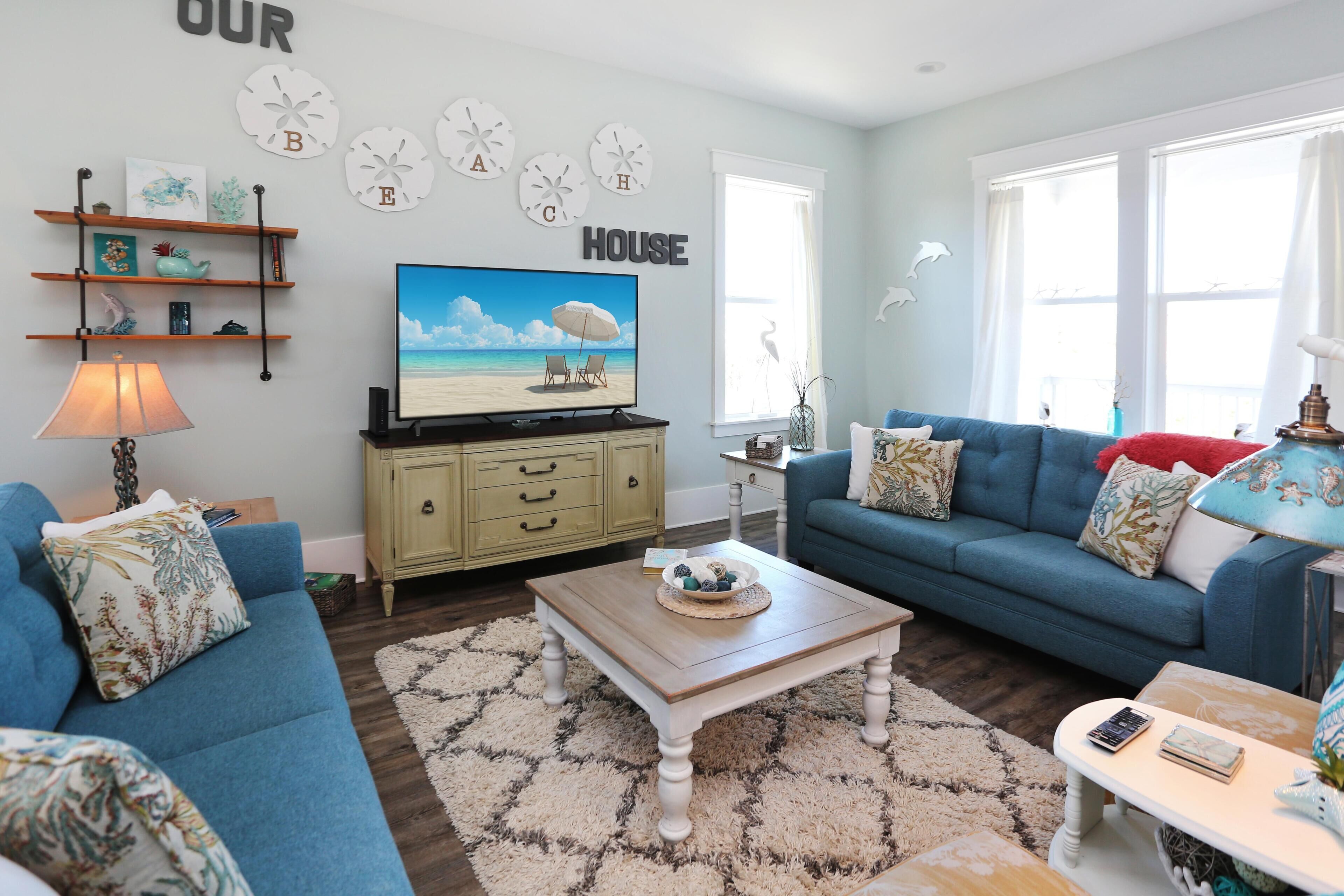 A living room with a blue sofa, a wooden coffee table, and a television displaying a beach scene. There are wall clocks and a sign that says "HOUSE".