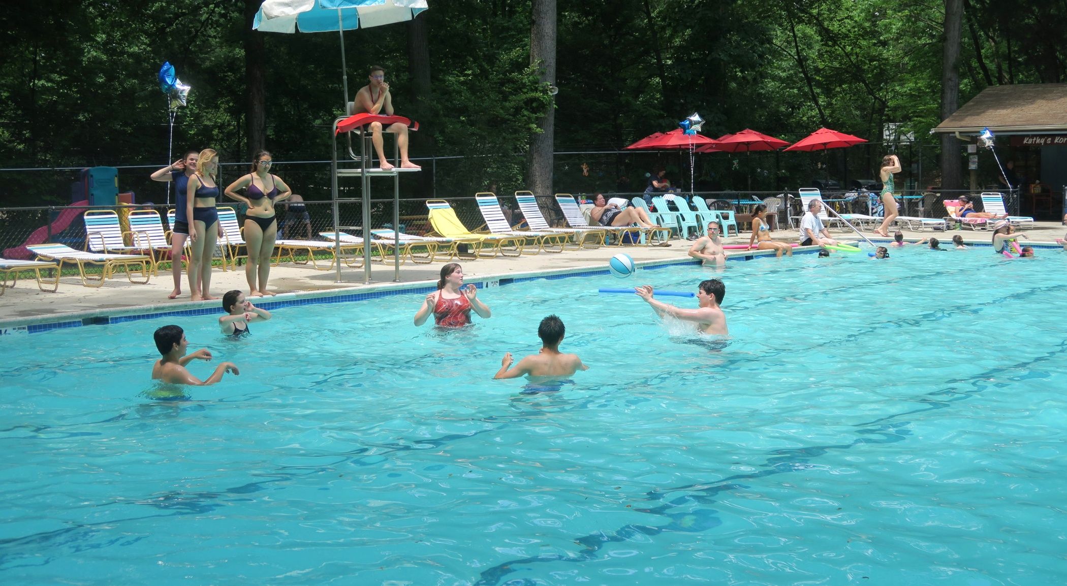 A swimming pool with people in the water, lounge chairs, and umbrellas around the pool area.