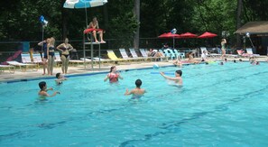 A swimming pool with people in the water, lounge chairs, and umbrellas around the pool area.