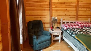 Una habitación tipo cabaña acogedora con techo de madera, una cama individual con manta escocesa, un sillón azul, una mesita de noche con lámpara y una ventana con cortinas.