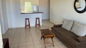 A living room with a brown sofa, a wooden coffee table, and two wooden stools. A kitchen area with a window and a hanging light fixture is visible in the background. - Beau T3 vue mer Proche Plage Wifi (Trois Ilets)