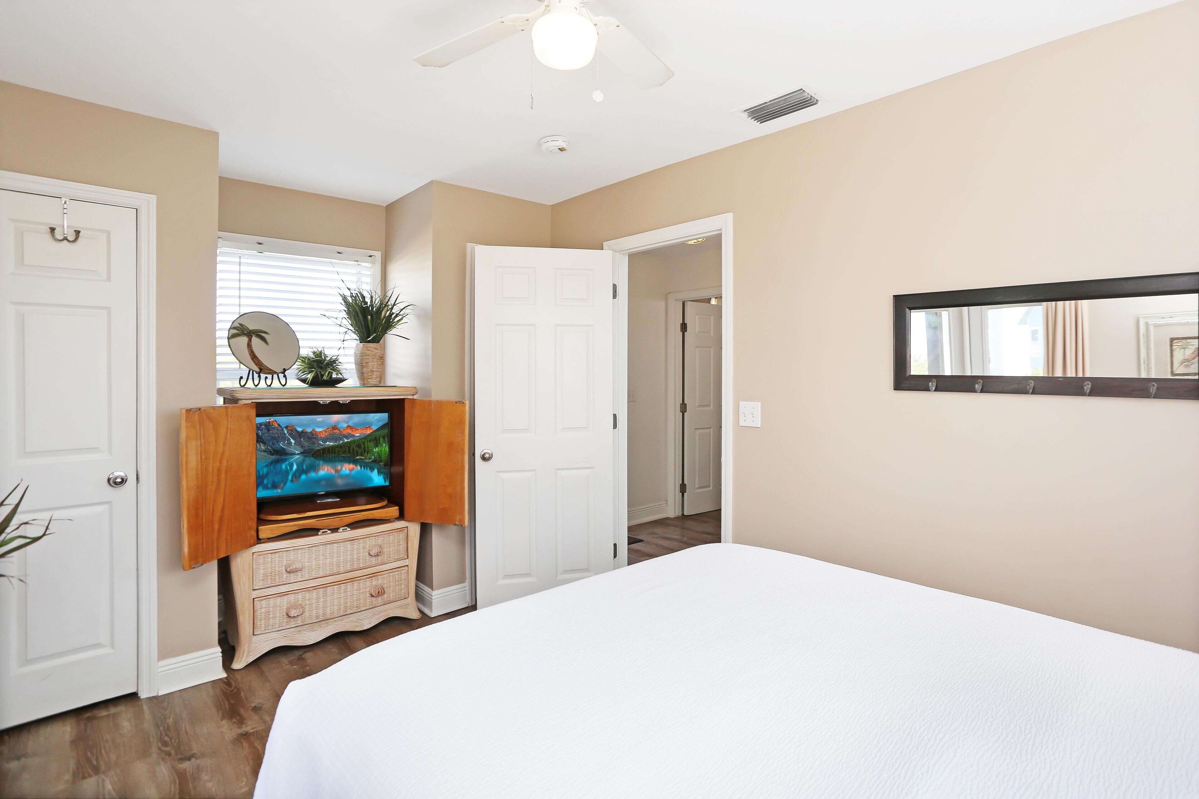 A bedroom with a bed, a television on a wooden stand, a mirror on the wall, and a door leading to another room.