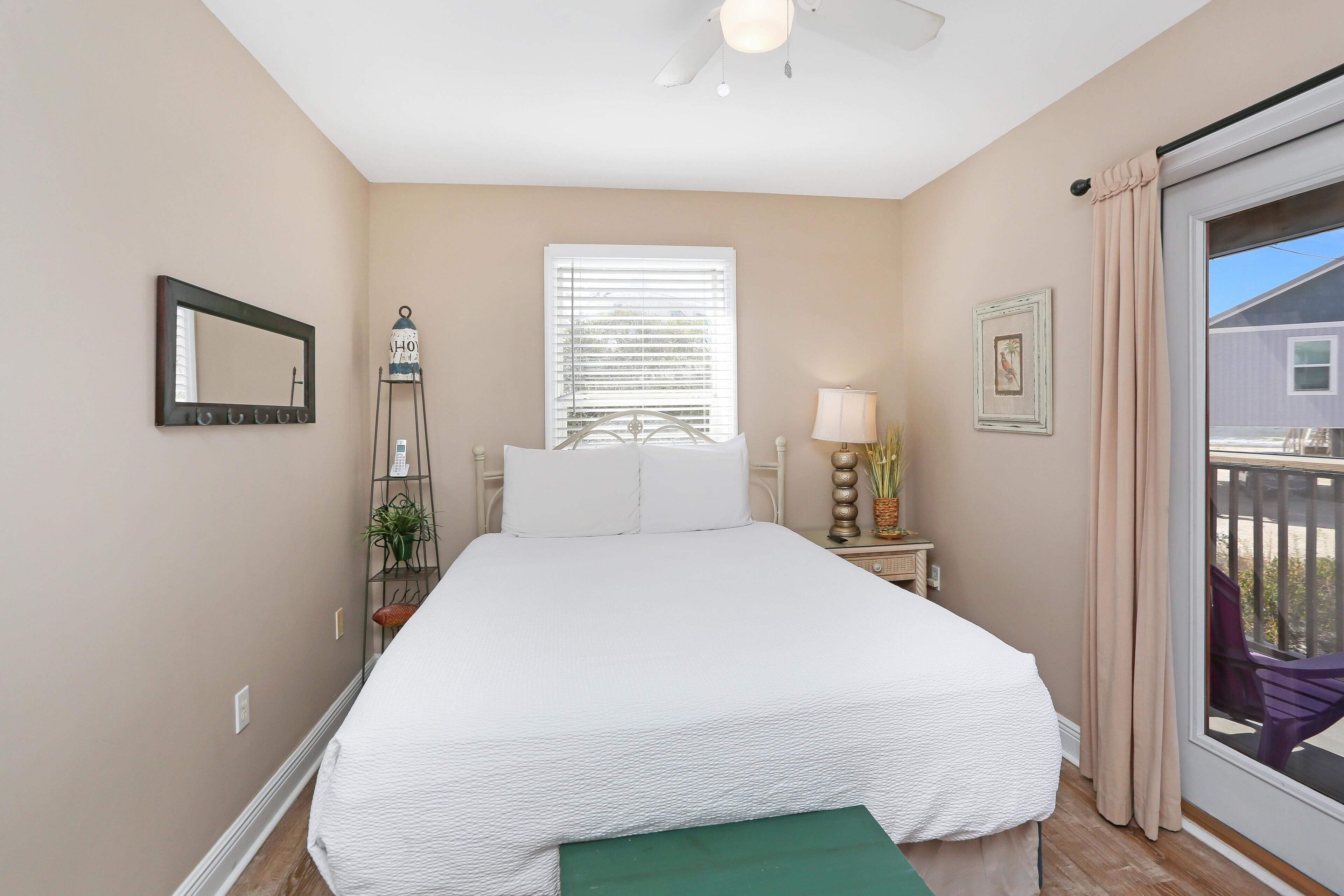 A bedroom with a large bed, a window with blinds, a bedside table with a lamp, a small plant, and a view of the outside balcony.