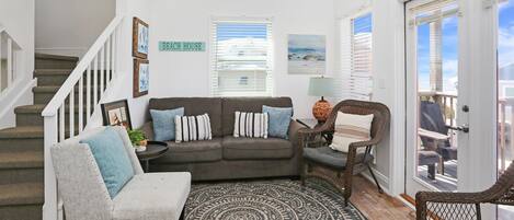 A living room with a gray sofa, a gray armchair, and a patterned rug. There is a staircase with white railings, a door labeled "Beach House," and a window with blinds.