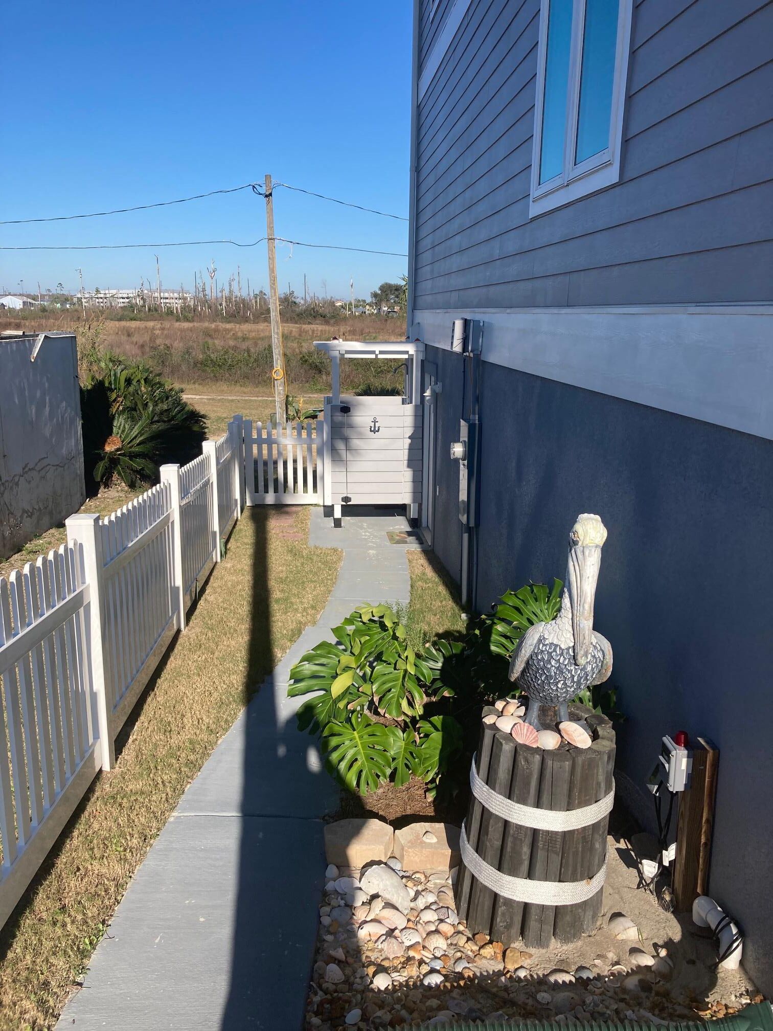 A narrow pathway leading to a house with a white picket fence, a wooden barrel with seashells, and a potted plant.