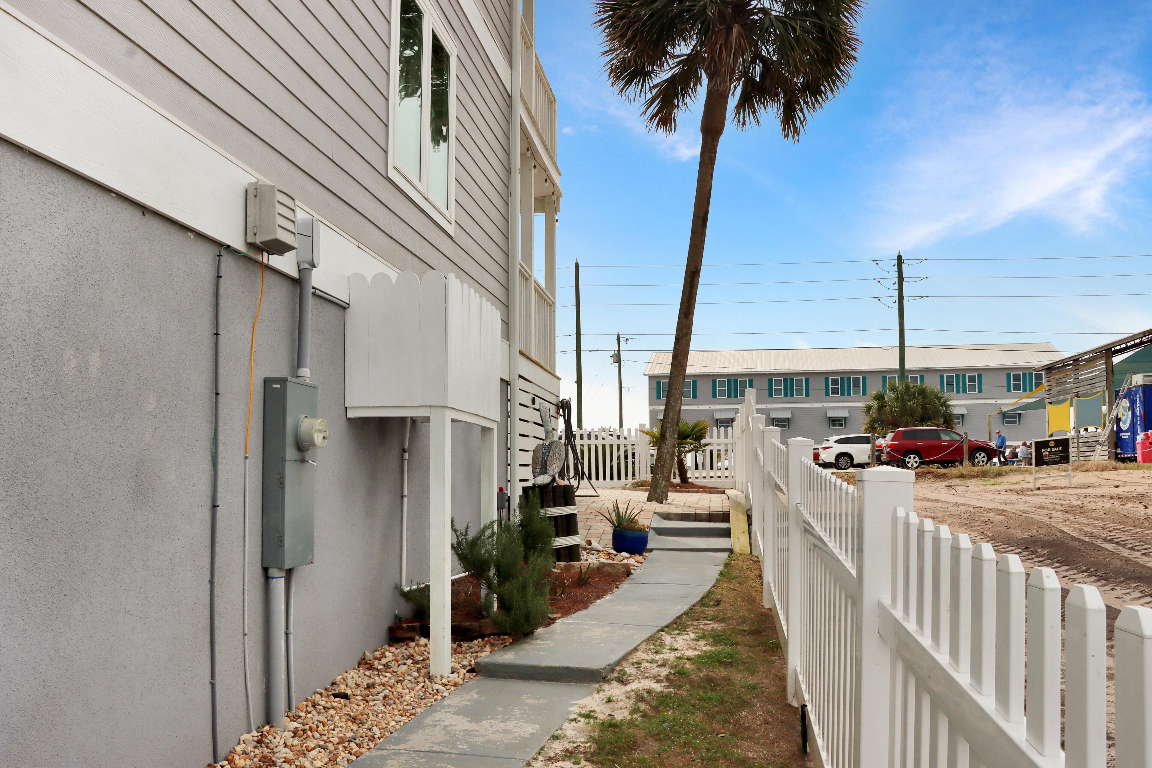 A narrow pathway between two buildings with a white picket fence, a palm tree, and a clear sky.