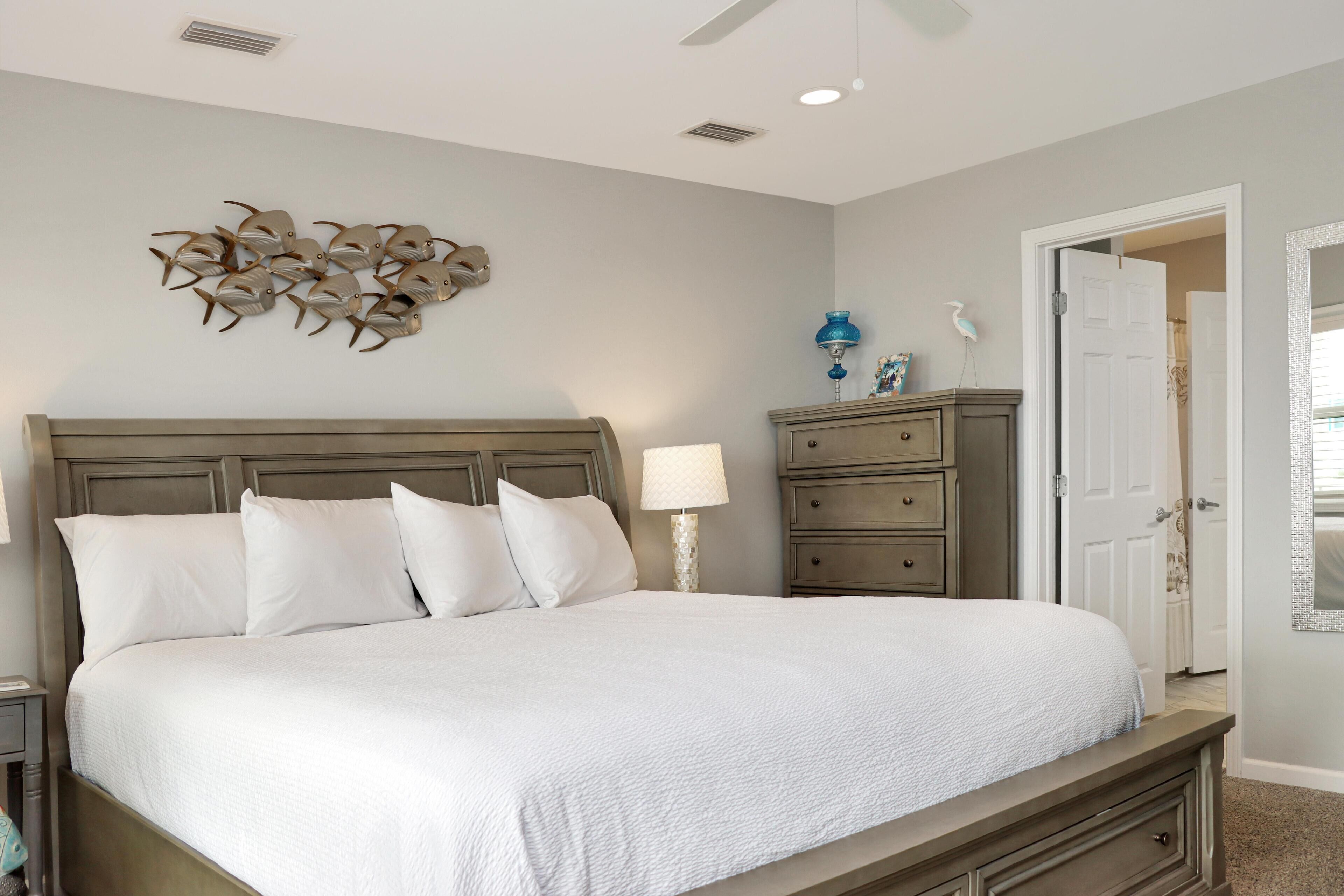 A bedroom with a large bed, a nightstand, a dresser, a wall-mounted lamp, and a decorative wall piece.