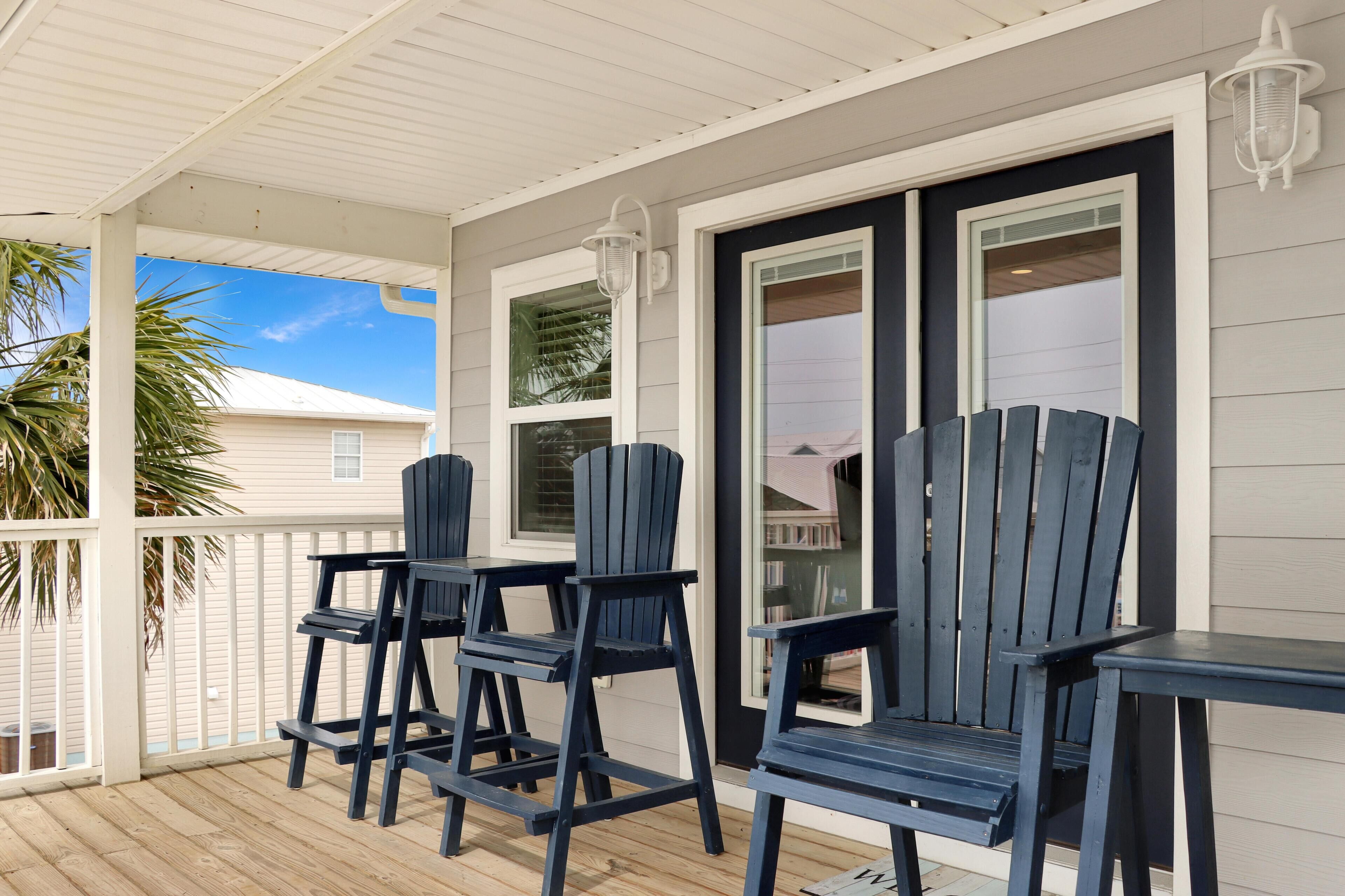 A covered porch with wooden flooring, three Adirondack chairs, and a table.