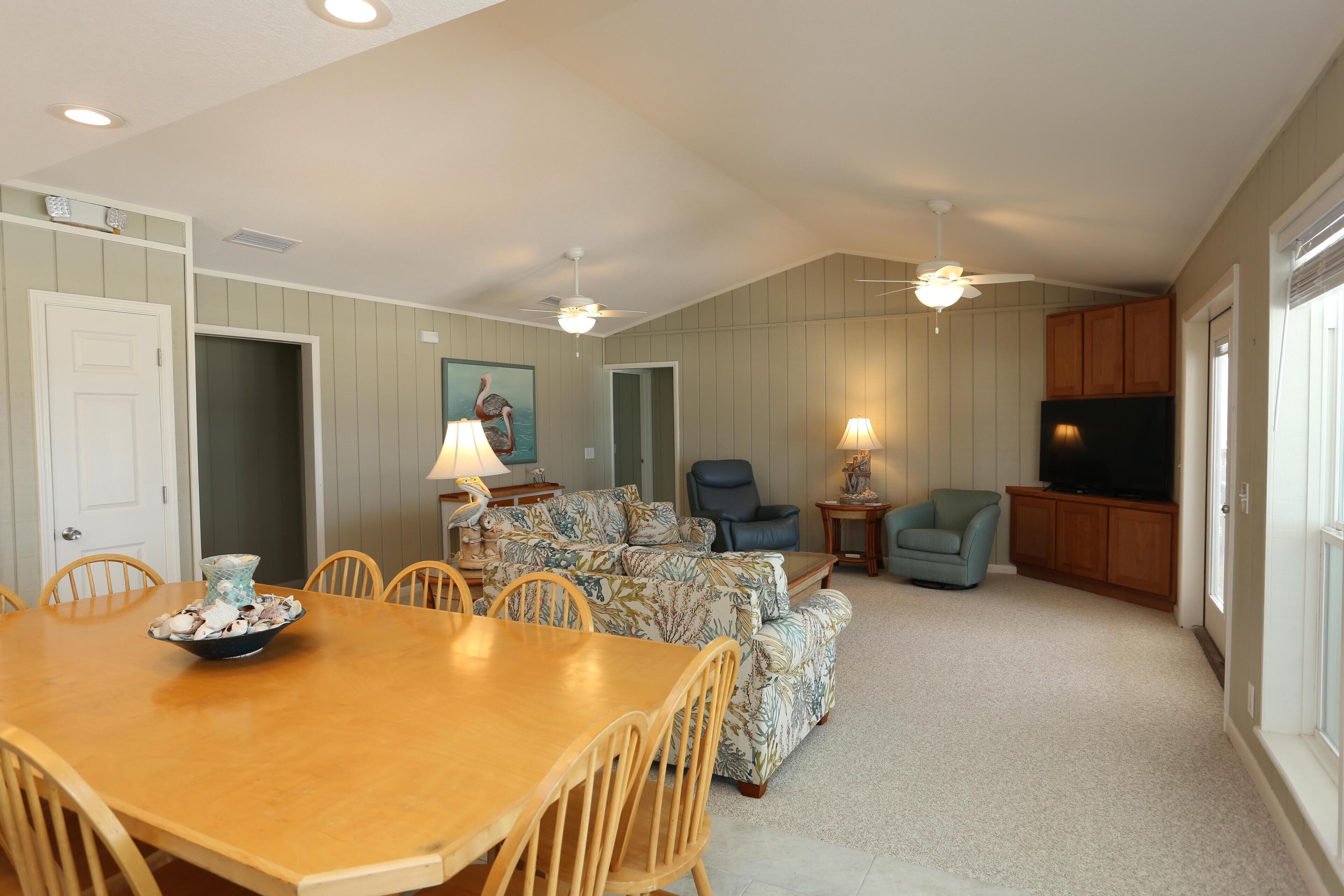 A spacious living room with a wooden dining table, comfortable seating, a television, and ceiling fans.