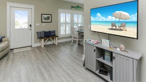Un salon avec un téléviseur diffusant une scène de plage, un coin repas avec des chaises et une bibliothèque remplie de livres et d’objets décoratifs.