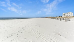 Une plage où l’on voit des empreintes de pas menant à une maison blanche, entourée d’une clôture et bordée de palmiers.