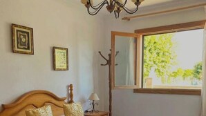 Un lit bien fait, avec une couette blanche, deux coussins dorés et un plaid décoratif. On y trouve une tête de lit en bois, une table de chevet avec une lampe et une fenêtre donnant sur des arbres.
