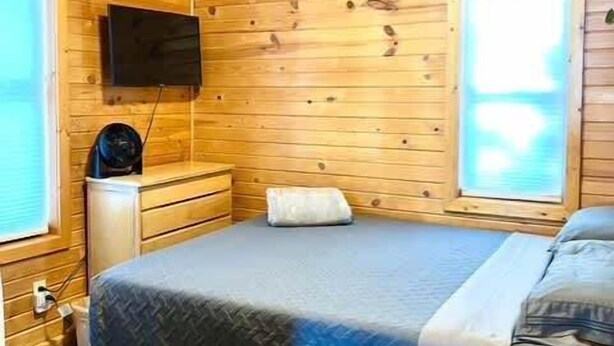 Una habitación de cabaña de madera con una cama, un aparador, un televisor montado en la pared y una ventana.