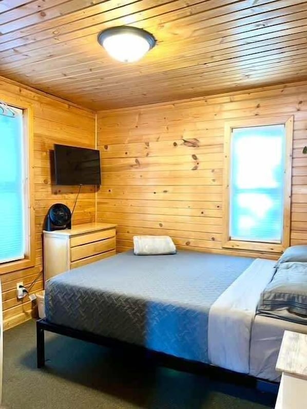 Una habitación de cabaña de madera con una cama, un aparador, un televisor montado en la pared y una ventana.