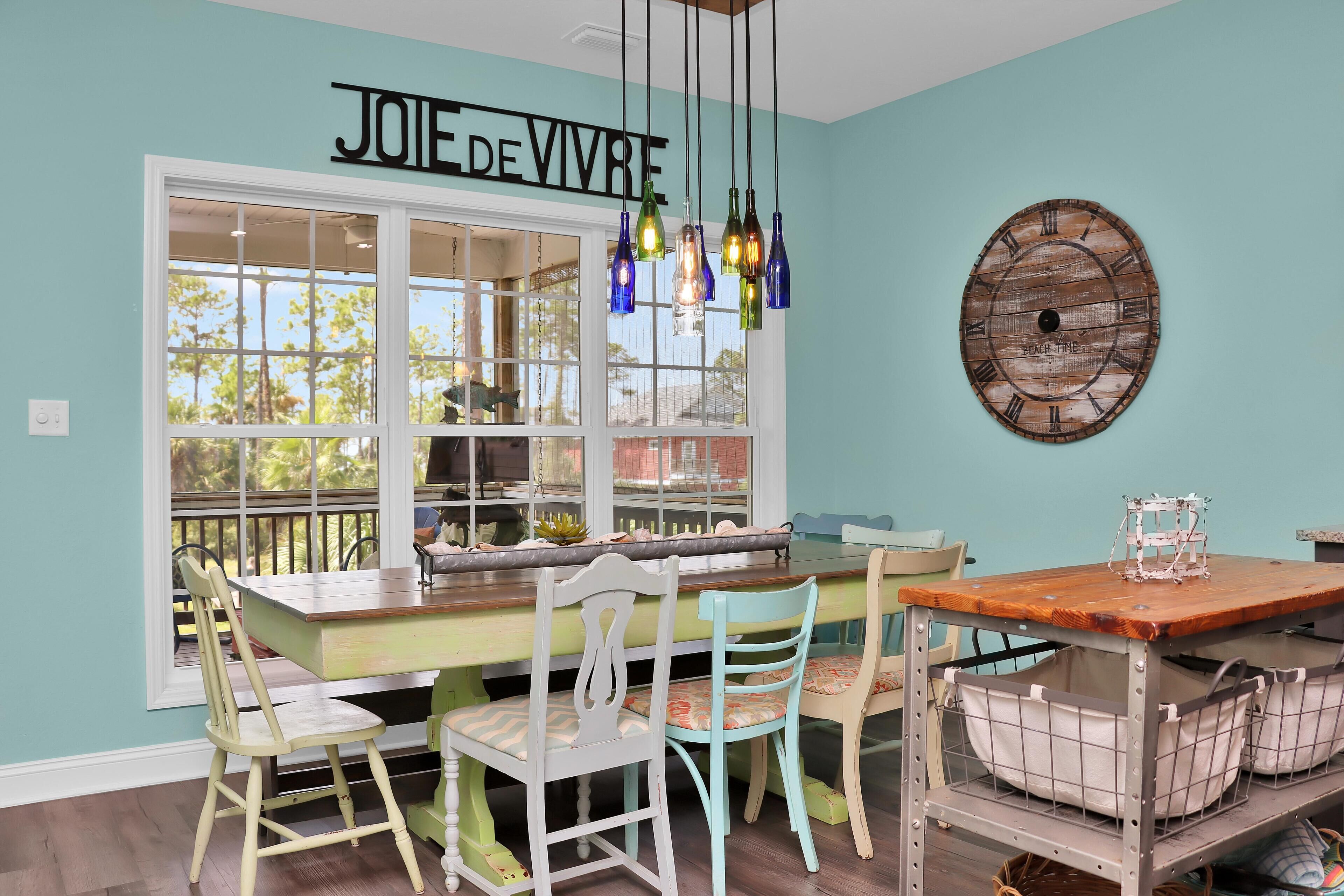 A dining area with a light blue wall, a wooden table, and chairs. There is a window with a view of trees and a sign above the window that reads "JOIE DE VIVRE".