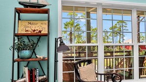 A cozy reading nook with a bookshelf, a chair, and a large window with a view of trees and a wooden deck. - Rehoboth Cottage (Port St. Joe)