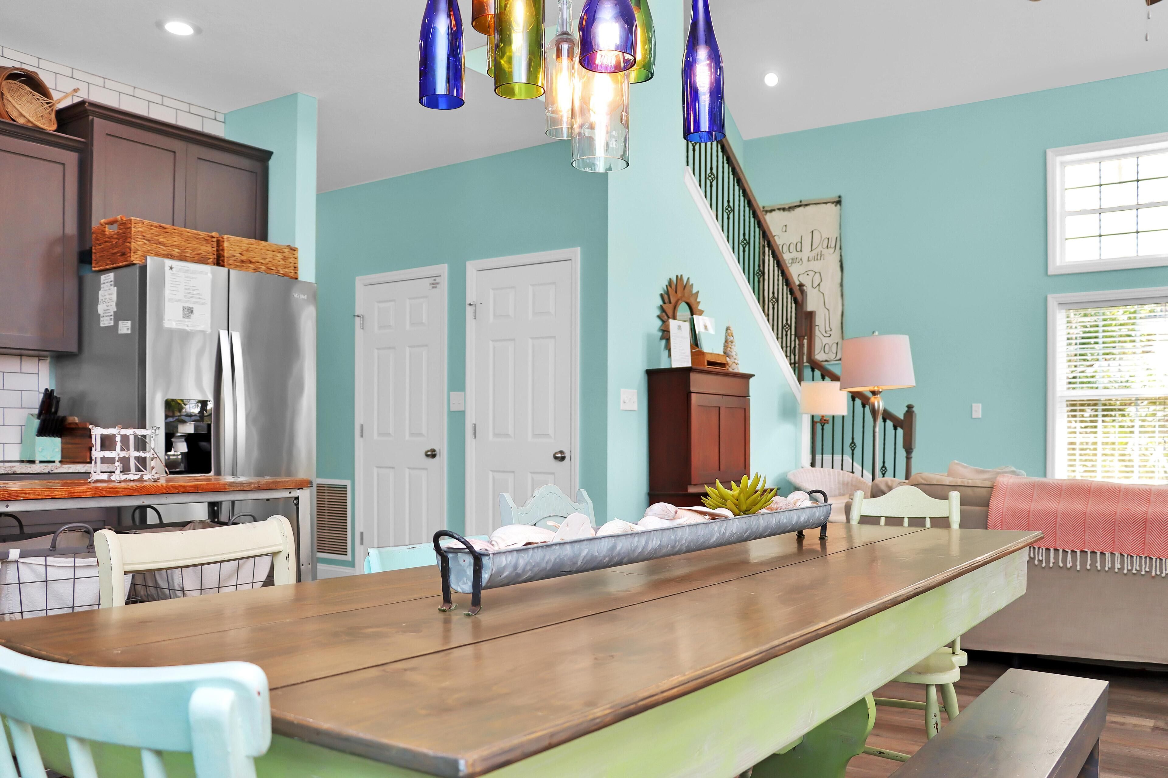 A kitchen with a wooden island, white chairs, and a dining table. There is a staircase leading to another level, and a window with blinds.