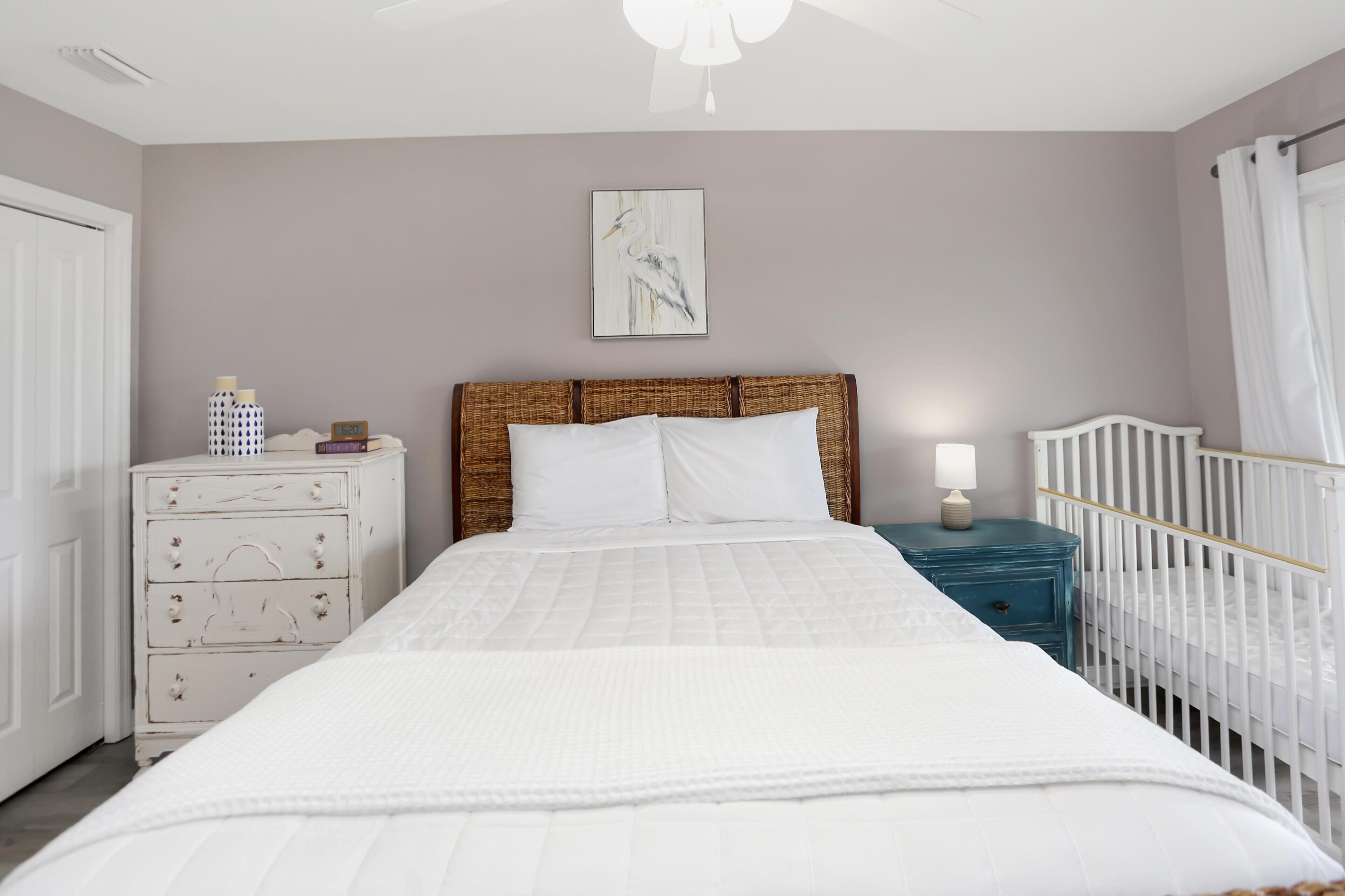 A bedroom with a bed, a nightstand, a dresser, a crib, and a wall-mounted picture.