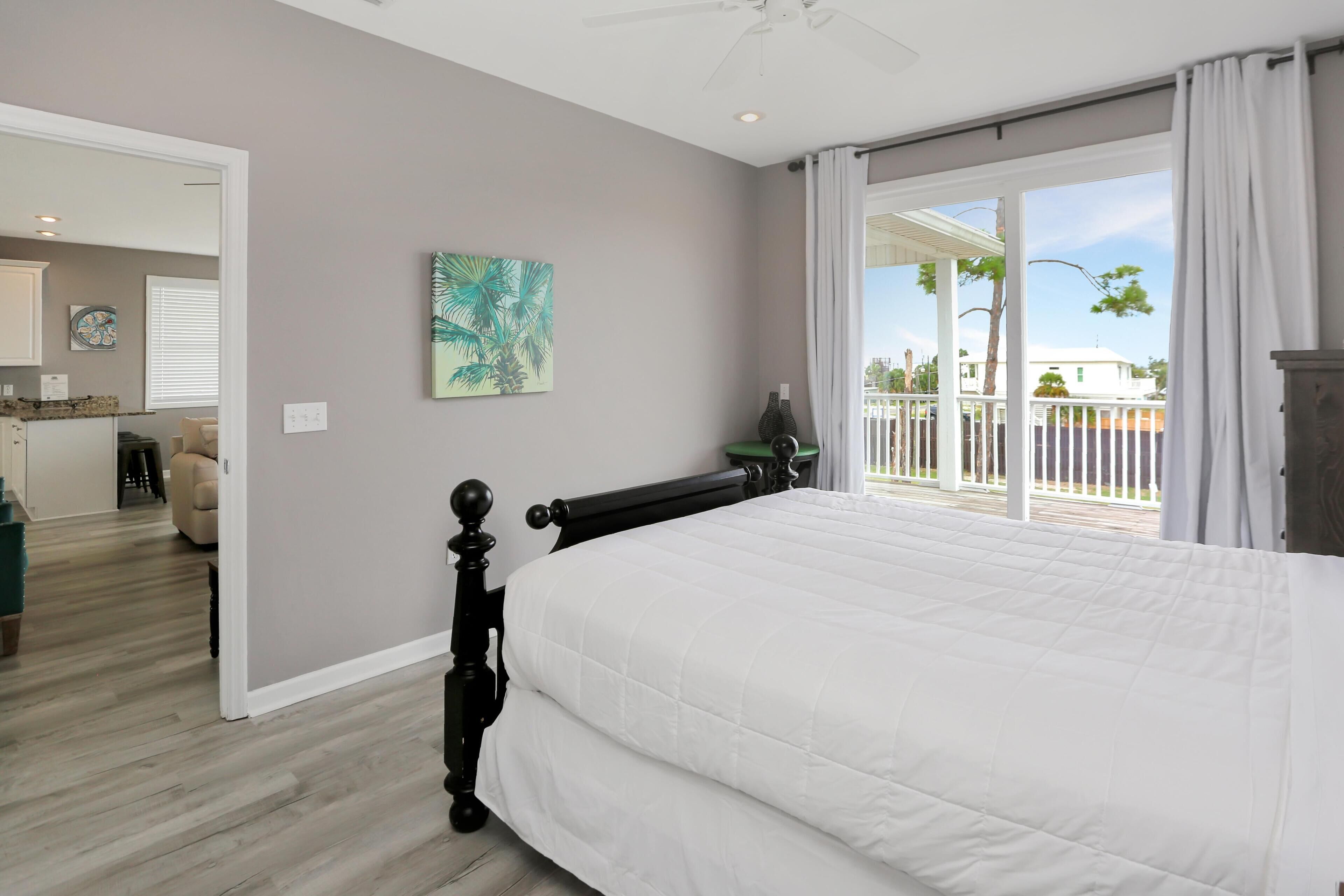 A bedroom with a large bed, a ceiling fan, a view to the outside, and a painting on the wall.