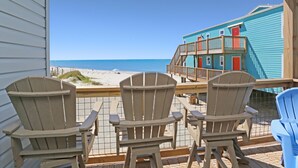A wooden deck with three Adirondack chairs facing a beach and a turquoise building with a balcony. - Paradise By The Gulf (Port St. Joe)