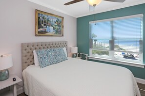 A bedroom with a tufted headboard, a large bed, a nightstand with a lamp, a window with blinds, and a ceiling fan. - Paradise By The Gulf (Port St. Joe)