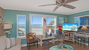 A living room with a teal wall, wooden flooring, a ceiling fan, a TV, a sofa, a dining table with chairs, and a small table with a lamp and decorative items. - Paradise By The Gulf (Port St. Joe)