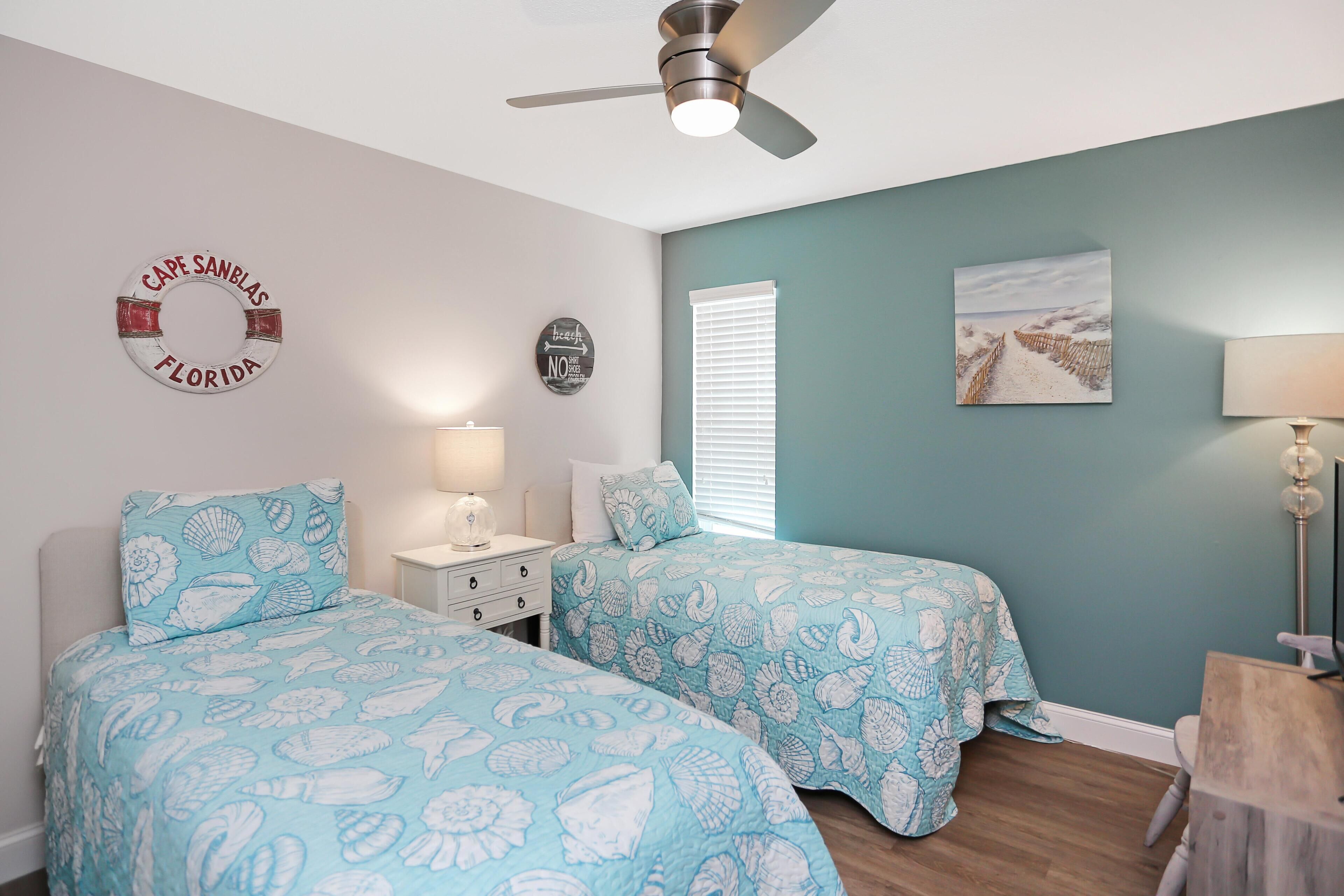 A bedroom with two beds, a ceiling fan, a nightstand, a lamp, and a beach-themed wall decor.