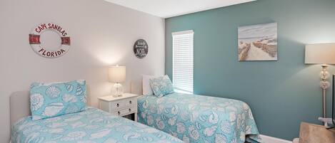 A bedroom with two beds, a ceiling fan, a nightstand, a lamp, and a beach-themed wall decor.