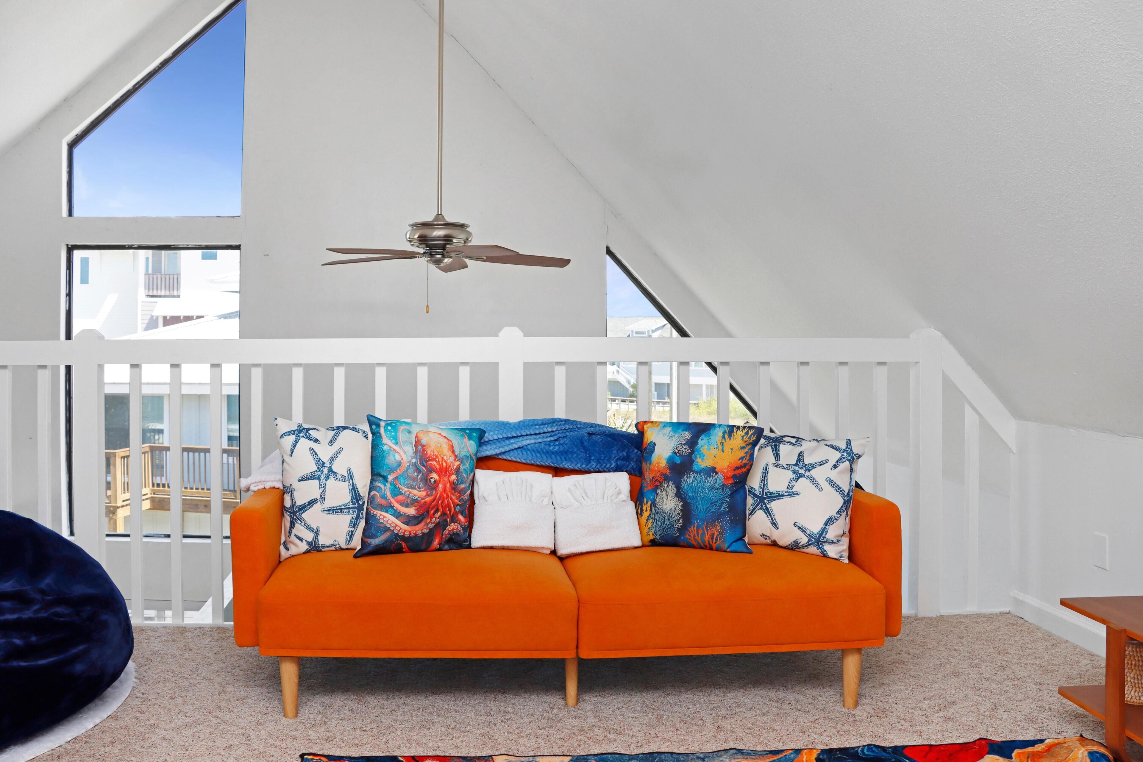 Une pièce avec un balcon blanc, un ventilateur de plafond et un canapé coloré agrémenté de coussins en forme d’étoiles de mer.