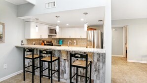 A kitchen with a bar area, white cabinets, a microwave, and a refrigerator. There are three bar stools and a countertop with a backsplash. - Sharkey's (Port St. Joe)