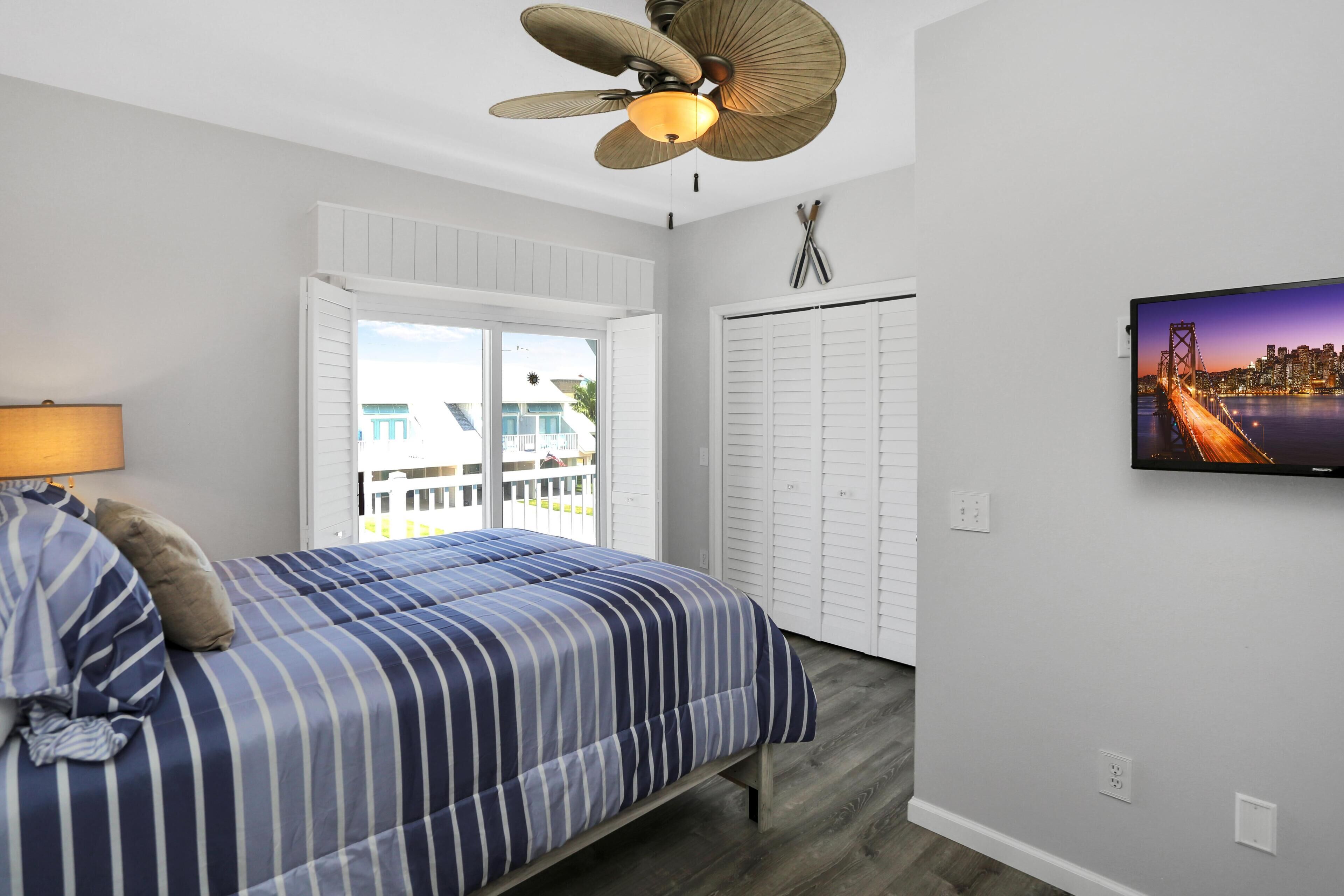 A bedroom with a bed, a ceiling fan, a TV, and a view of a cityscape through a window.