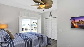 A bedroom with a bed, a ceiling fan, a TV, and a view of a cityscape through a window.