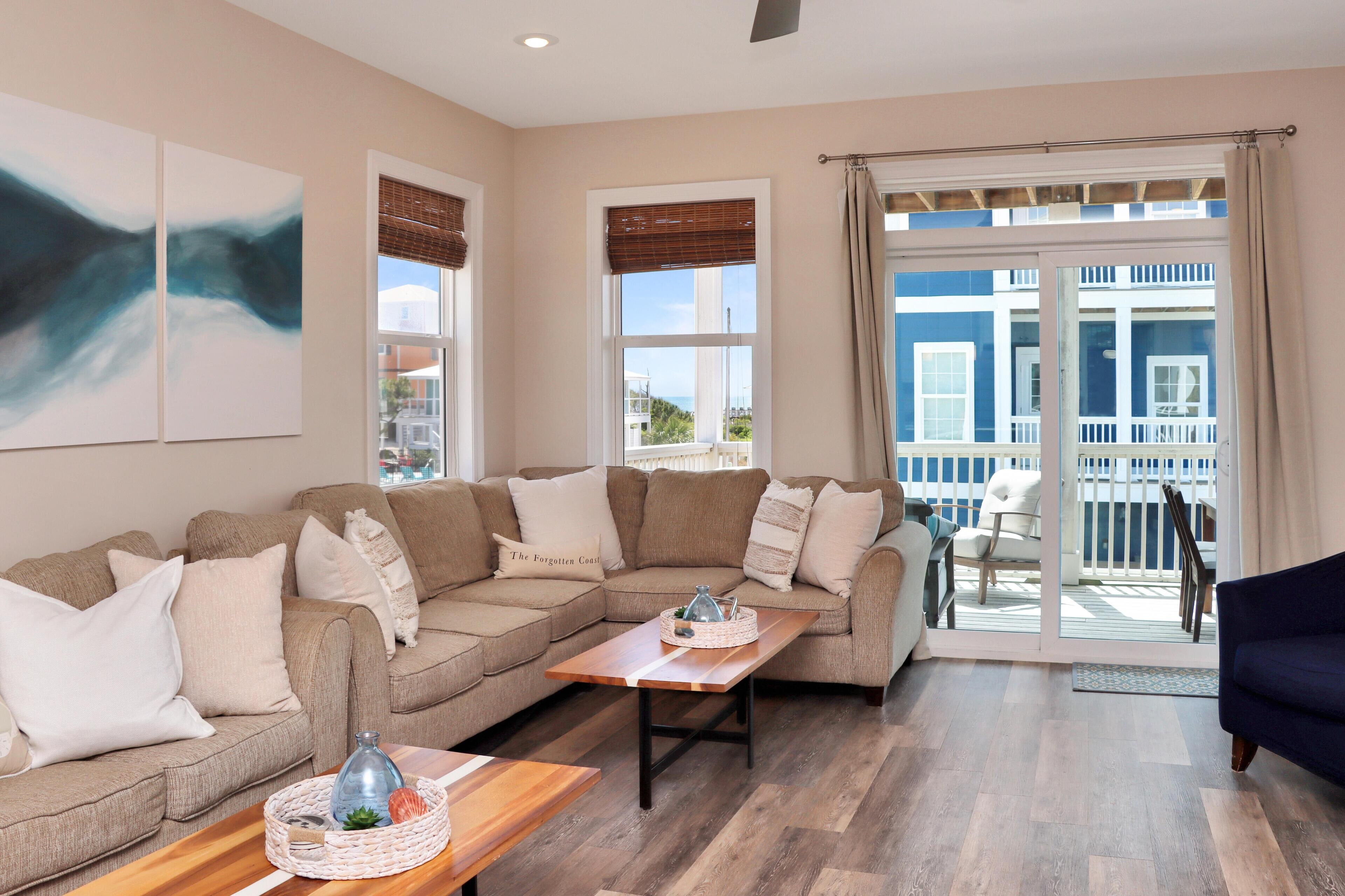 Un salon avec un canapé d’angle beige, une table basse en bois et une vue sur un balcon aménagé avec du mobilier d’extérieur.