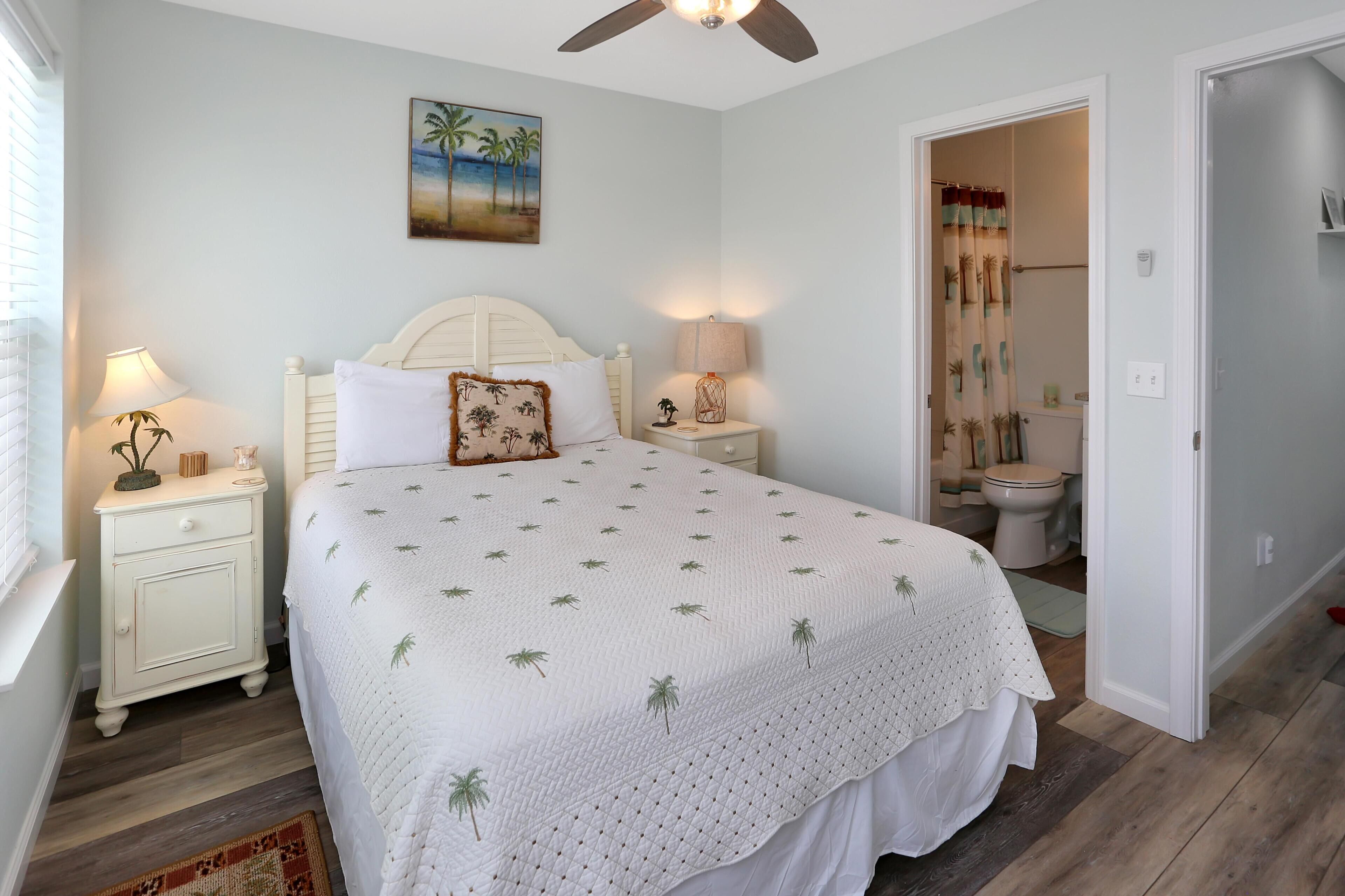 A bedroom with a bed, nightstands, a ceiling fan, a painting, and a bathroom visible through an open door.